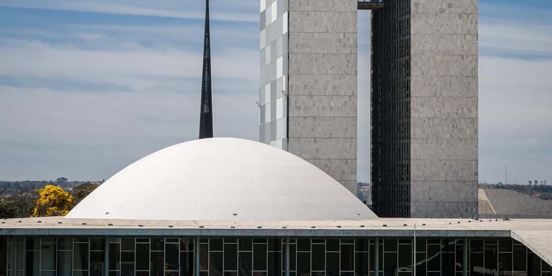 Aerial view of Brazil's National Congress building with its twin towers and dome in Brasília, with the national flag flying beside it.