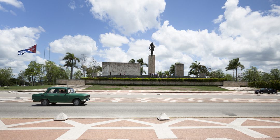 A vintage green car driving past a monument with the Cuban flag flying nearby.