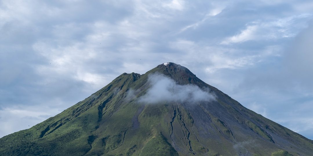 Symmetrical volcano cone rising above green forested slopes under a cloudy sky