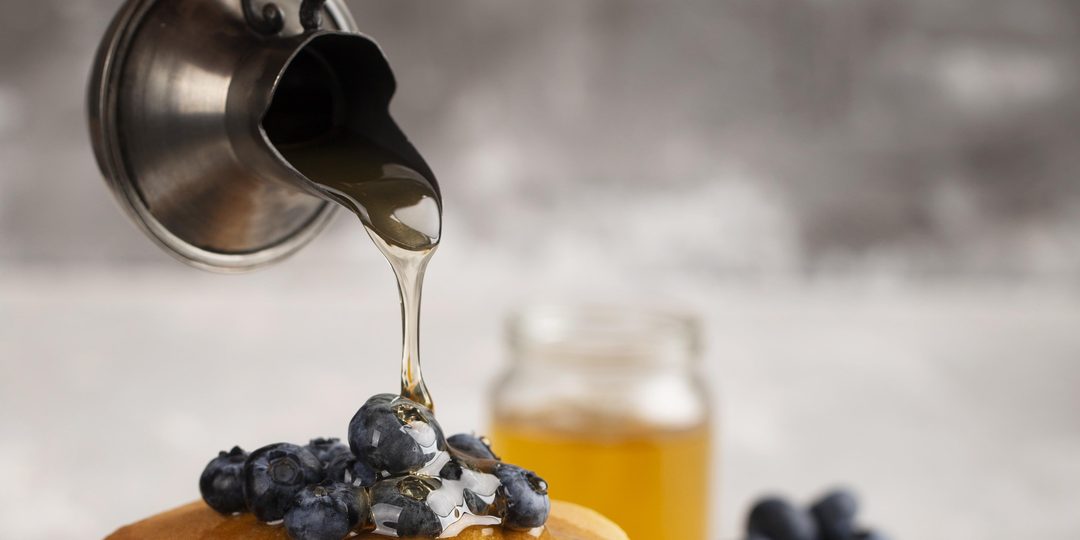 Maple syrup being poured from a small tin over a stack of pancakes topped with blueberries.