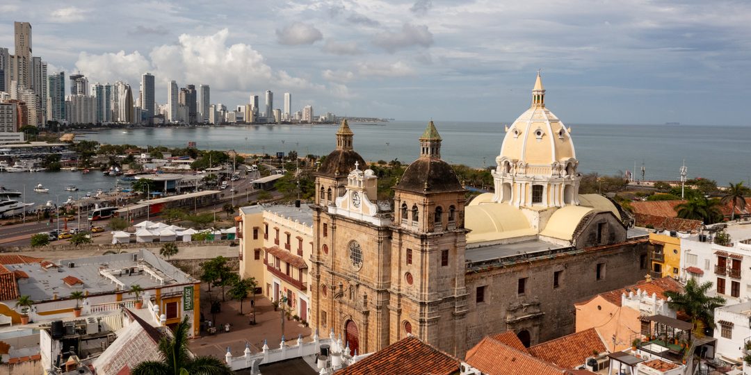 Aerial view of the domed San Pedro Claver Sanctuary in Cartagena's historic walled city, with the modern skyline and Caribbean Sea behind it.